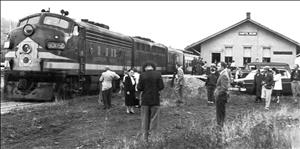 People stand in field next to a diesel locomotive and a small wooden depot building. 